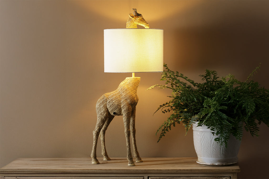 textured golden giraffe lamp with head poking out from top of white fabric shade set on wooden dresser next to fern plant