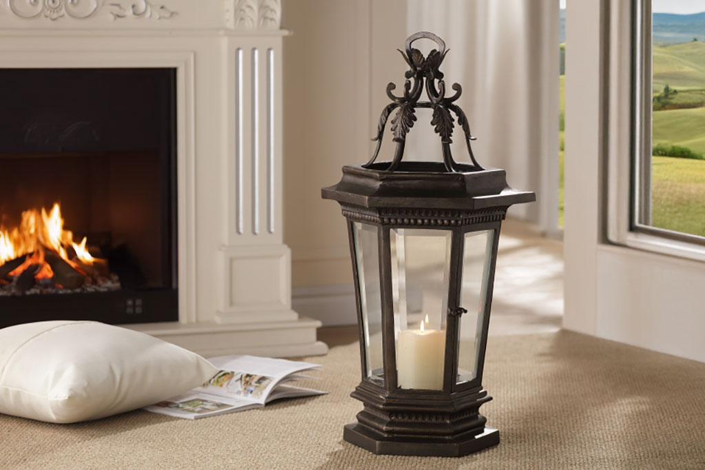 Acanthus garden lantern shown in home. It sits on the floor near a lit fireplace with view of countryside seen through French Doors