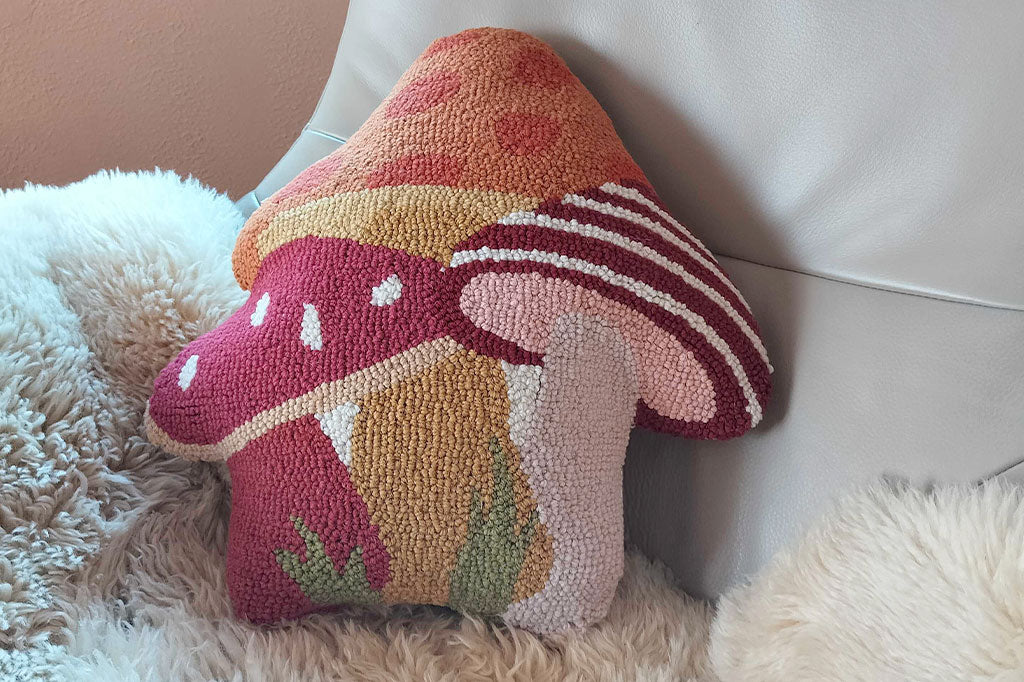mushroom shaped pillow, cluster of three mushrooms in the colors of orange, red, yellow, and white. Shown on easy chair with fleece blanket