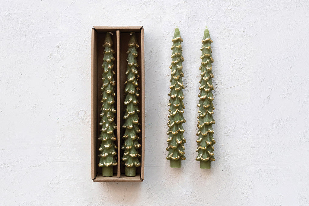 Two pine tree-shaped candles in a brown rectangular box, with two additional candles standing beside it on a white textured background.