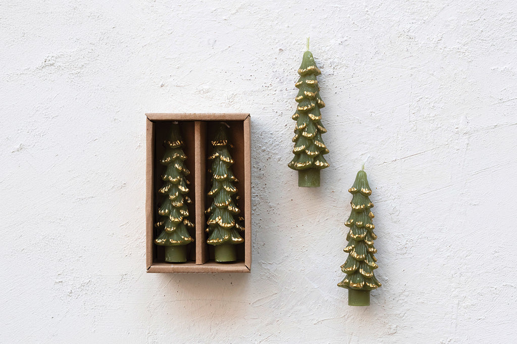 Four olive green Christmas tree-shaped candles, with two standing outside in a wooden box and one standing outside to the right, set against a textured white wall.