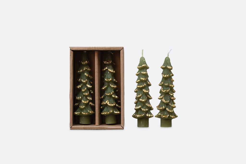 Four Christmas tree-shaped candles in olive green with gold glittery accents, shown with two inside a brown cardboard box and one standing outside beside it on a light gray background.