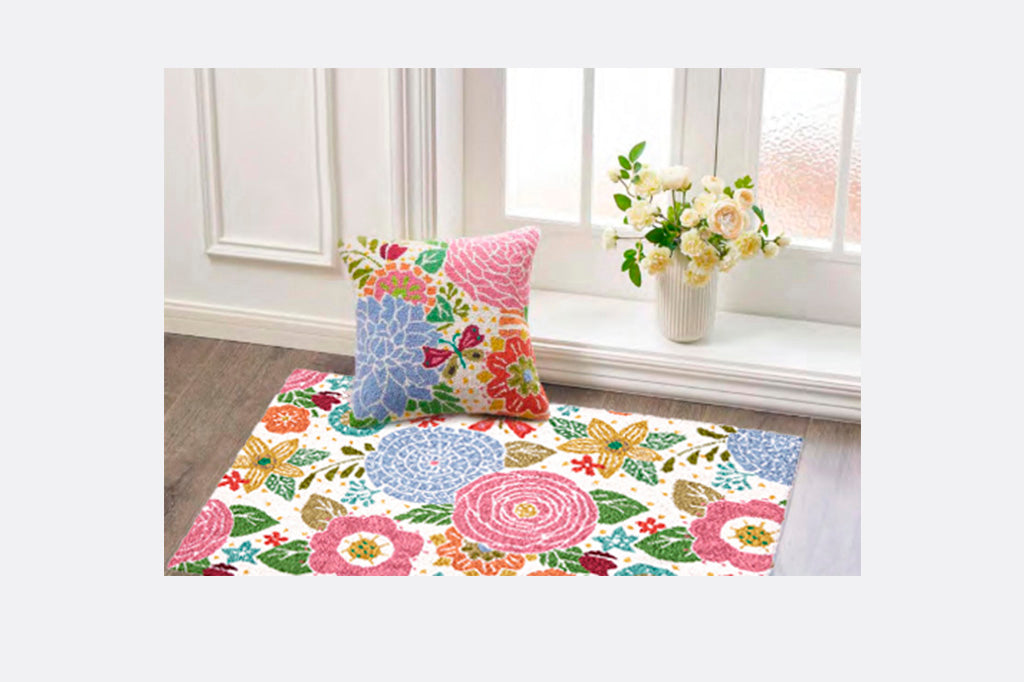 Hooked wool throw rug shown on floor next to coordinated throw pillow in a bright room by vase of flowers; scattered floral print in playful shades of pink and blue