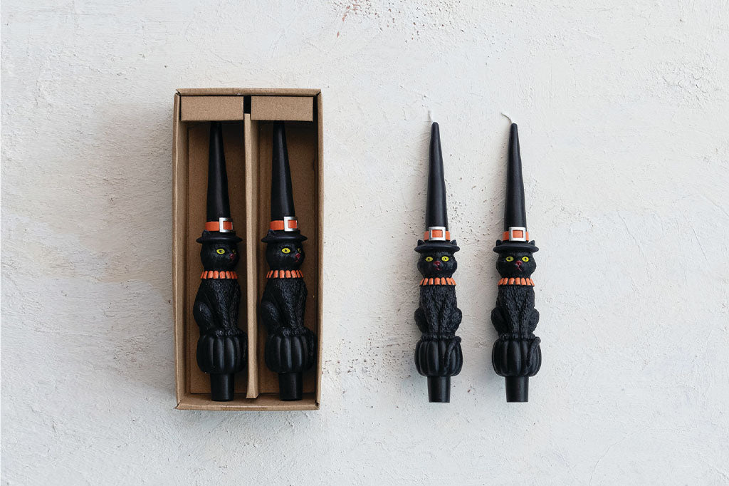 four taper candles, each featuring a black cat in a classic witch hat with bright orange details