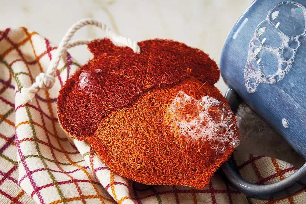 Red and orange scrubber with soap suds next to a blue mug on a checkered towel