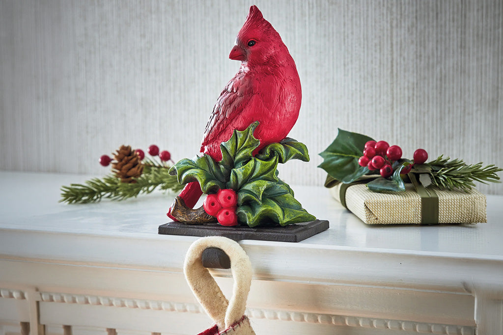 A red cardinal sculptured stocking hook perched on green holly with red berries, displayed on a white mantel alongside holiday greenery, pinecones, and a wrapped gift box.