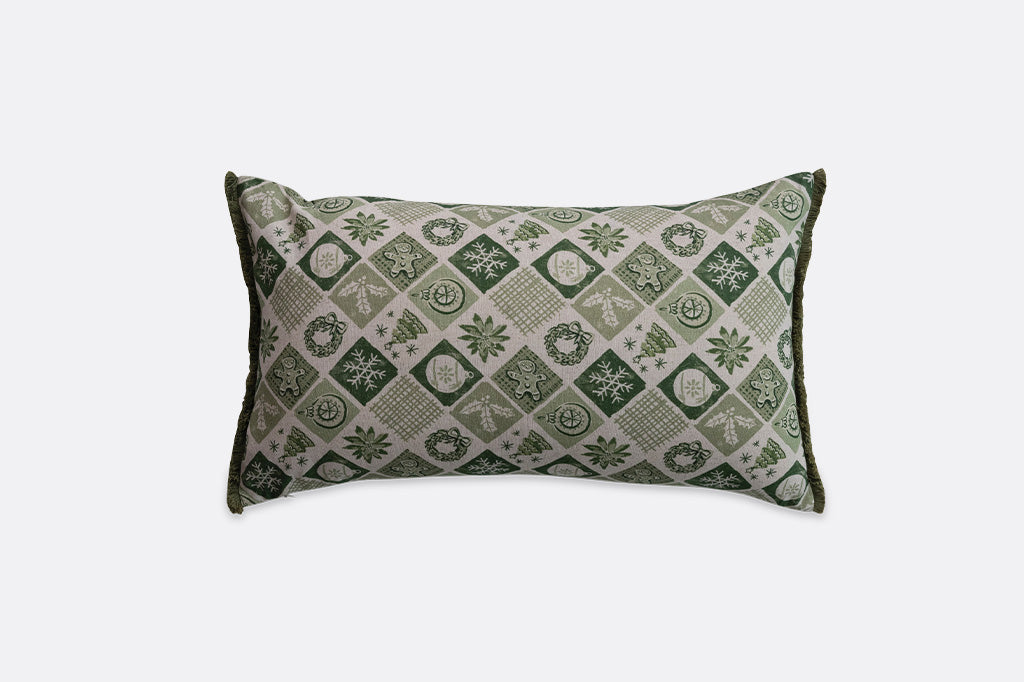 A rectangular lumbar throw pillow featuring a green and beige geometric diamond pattern with festive motifs like snowflakes and wreaths, set against a plain light gray background.