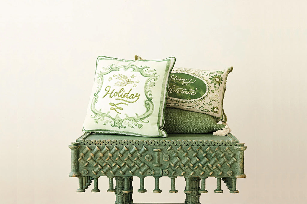 Two decorative throw pillows styled on a distressed green wooden bench against a plain off-white background. One pillow is light green with green ornate embroidery reading 'Holiday', and the other is cream with green embroidery reading 'Merry Christmas' and a green oval patch.