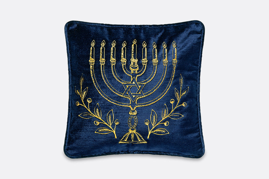 A square navy blue velvet pillow featuring a gold embroidered menorah, Star of David, and botanical motifs on the front, set against a light gray background.