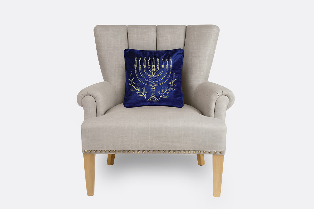 A light beige upholstered wingback armchair with wooden legs, featuring a dark blue velvet throw pillow with a gold embroidered menorah design, set against a plain light gray background.
