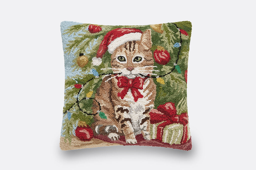 light blue square decorative throw pillow with a tabby cat wearing a santa hat and red bow in front of a christmas tree and gifts entangled in christmas lights, all pictured on grey background