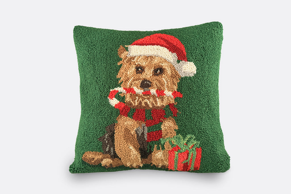 Square green decorative hooked pillow with yorkie pictured on pillow holding candy cane in its mouth with santa hat on and winter scarf, with a gift by its side pictured on light grey background