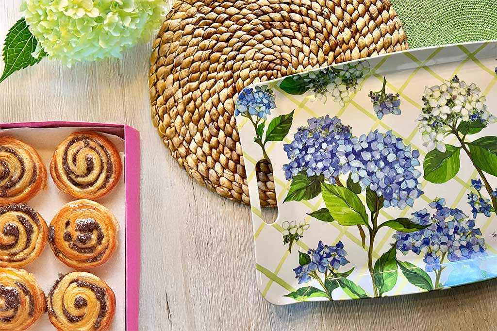wood serving tray with printed design of green lattice and hydrangea shown on a table with scattered items, top view. A box of donuts, straw placements, and a bouquet of white hydrangeas.