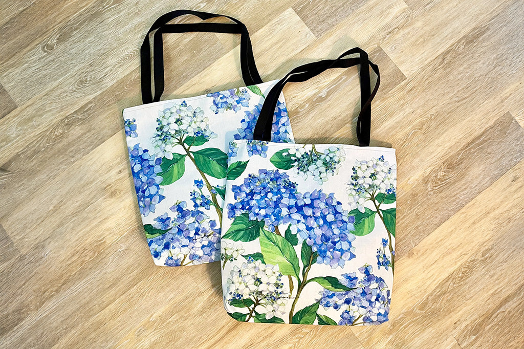 Two tote bags shown on wood floor featuring garden print of blue hydrangeas. Black handles. Sold individually.