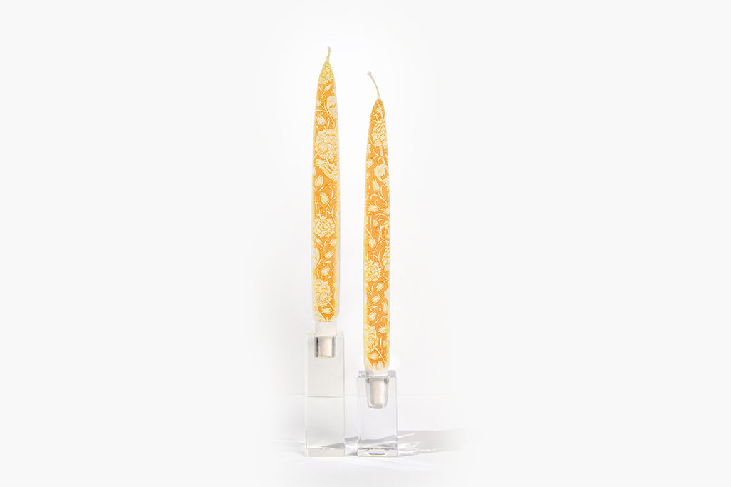 Pair of flat, taper candles with decorative painted floral motif in orange, displayed in glass candle holders on a white background