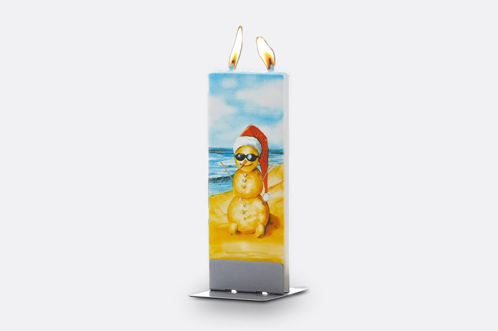 A tall rectangular candle with two lit wicks, featuring a printed illustration of a snowman wearing a Santa hat and sunglasses on a beach. The candle has a silver metal base and is photographed against a plain light gray background.