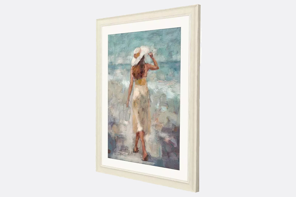 side view of an impressionistic beach scene captures a graceful woman walking along the beach. Matted in buff and framed in a distressed ivory wood.