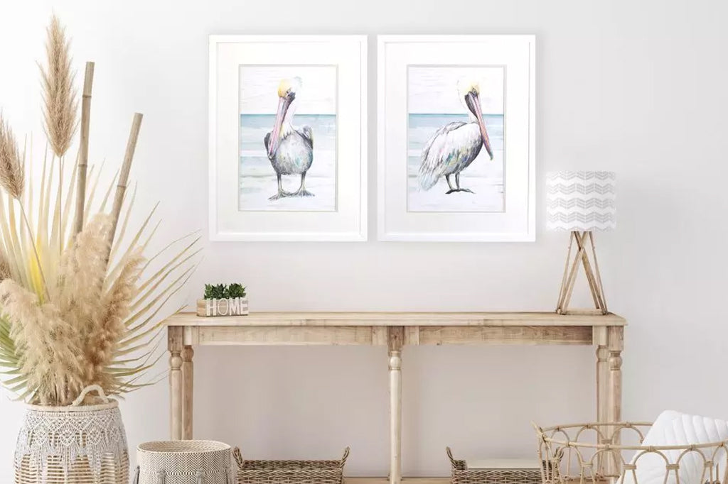 A set of two pelicans walk along the beach. Prints are double matted with a white top mat and framed in a modern flat white moulding. Mounted on wall above console table