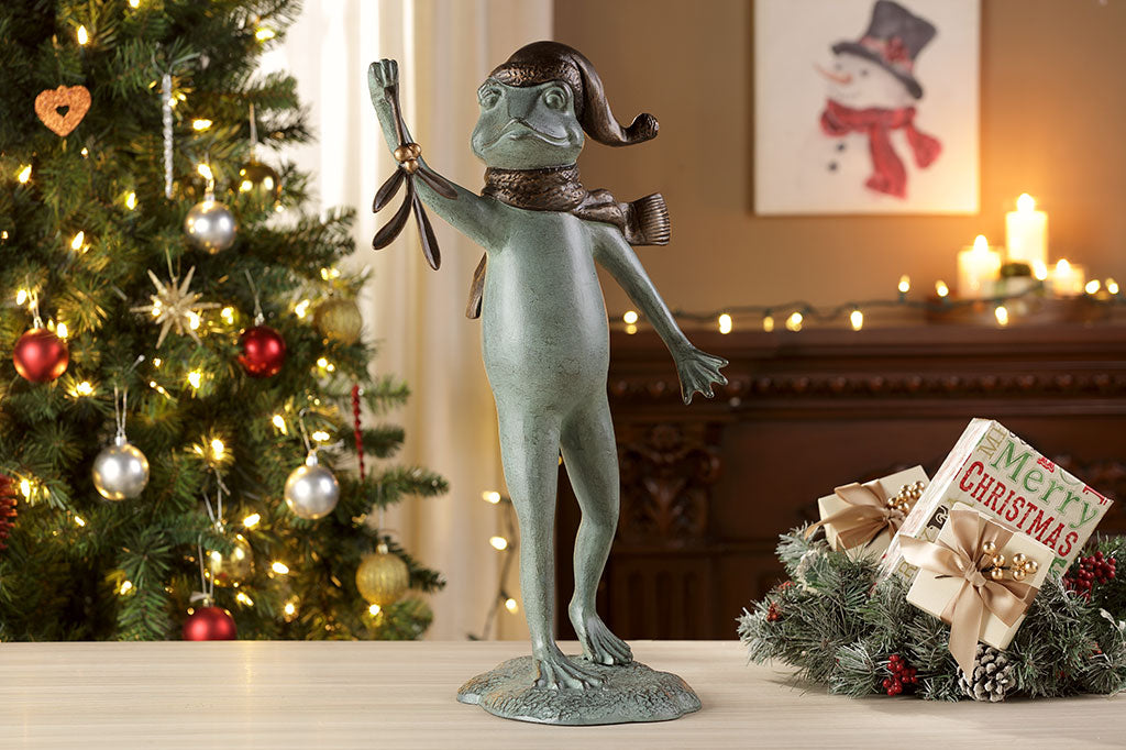 garden sculpture of frog in winter garb holding mistletoe from outstretched webbed hand. It stands upon an indoor table with Christmas tree in background and holiday decor in background.