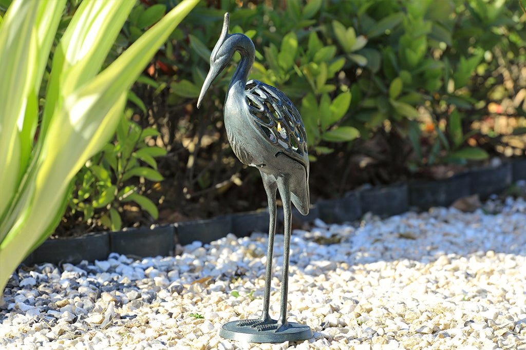 Modern metal heron candleholder with white lit candle face left placed in outdoor garden space