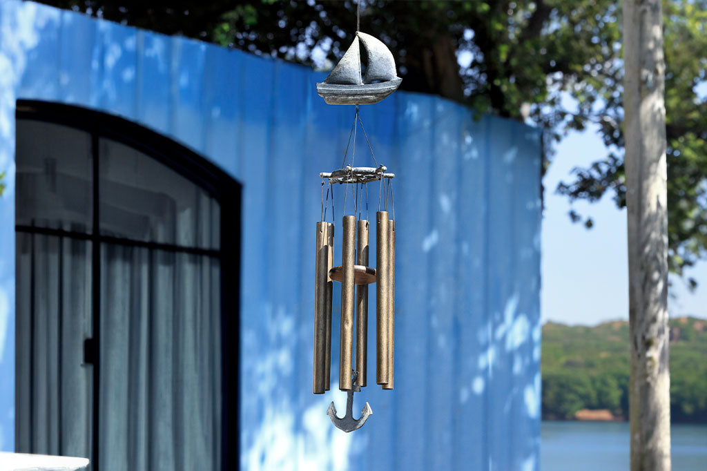 Wind chime with cast metal sail boat motif in zinc finish has 6 tube chimes and a pendant shaped like an anchor, hanging from lakeside tree near rustic boat house