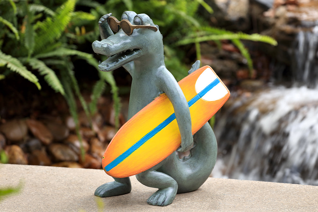 alligator garden sculpture with sunglasses holding an orange and yellow surf board 