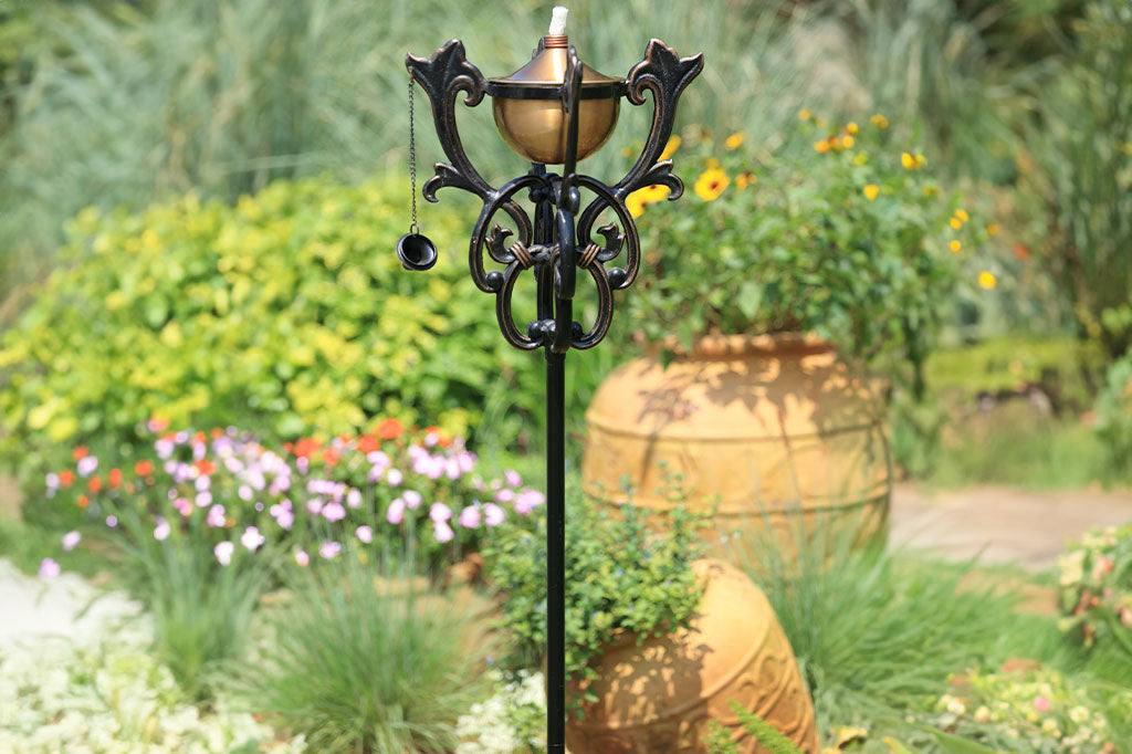 scrollwork encases the oil reservoir on a staked garden oil torch shown in a flower garden