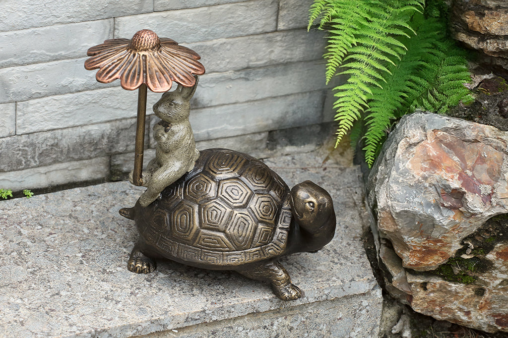 Bronze turtle with rabbit under a flower-shaped umbrella on a stone surface with ferns and rocks.