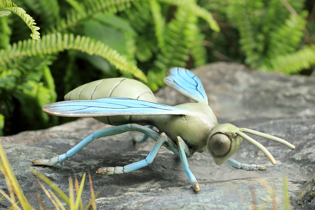 Sculpted metal wasp in soft shades of blue and sage green shown on a rock in a fern garden