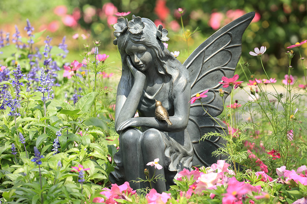 cast aluminum garden sculpture of a sitting fairy adorned with a flower crown and a little bird sitting on her arm. Placed in garden bed surrounded by purple and pink blooms