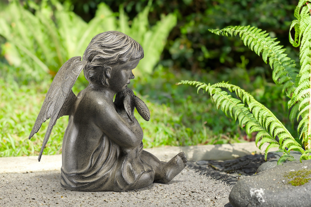Cast metal cherub with wings holding bunny in blanket, sitting in a fern garden, facing right