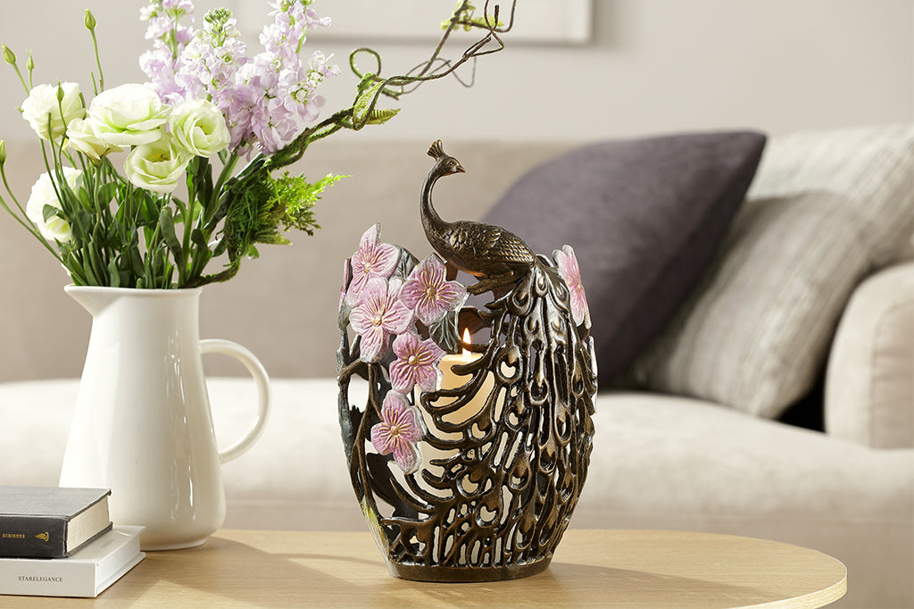 cast metal candleholder with peacock feathers and blossoms holding white pillar candle set on coffee table