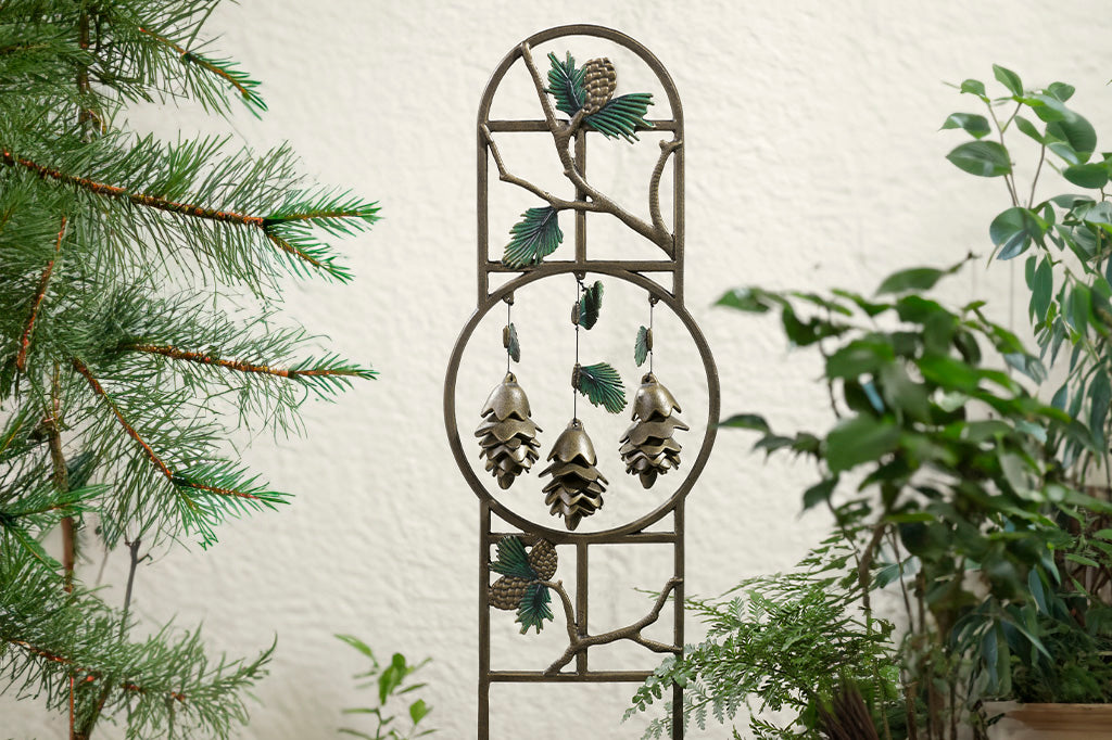 Pinetree theme garden trellis with 3 pinecone shaped pendants hanging within sculptural hoop. Trellis is shown against stucco wall in outdoor scene with pine branches and potted plants