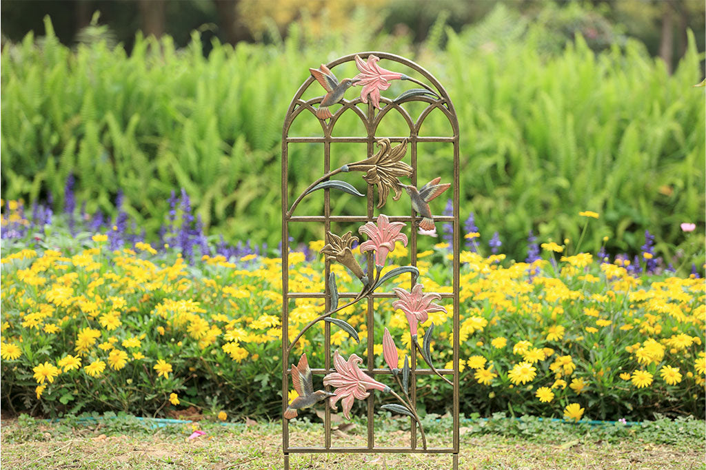 arched trellis featuring pink and gold lilies with sculpted hummingbirds at 3 blossoms. Shown in blooming garden with ferns, purple larkspur and yellow daisies