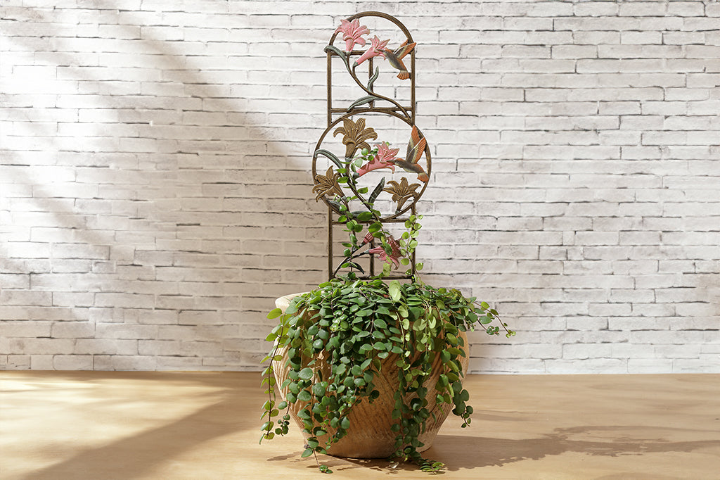 pink and bronze planter trellis with lilies and hummingbird details. Trellis placed in garden pot with trailing vine plant