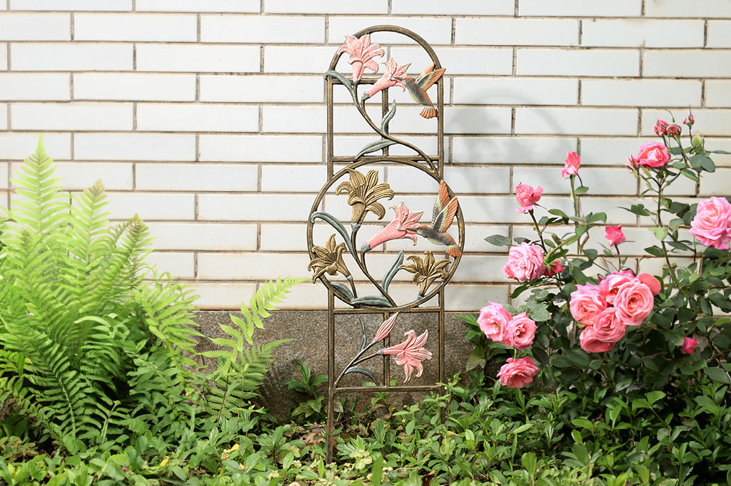 Cast metal planter trellis with lilies and hummingbird motifs and circle detail in the center. Trellis placed in garden bed besides outdoor wall