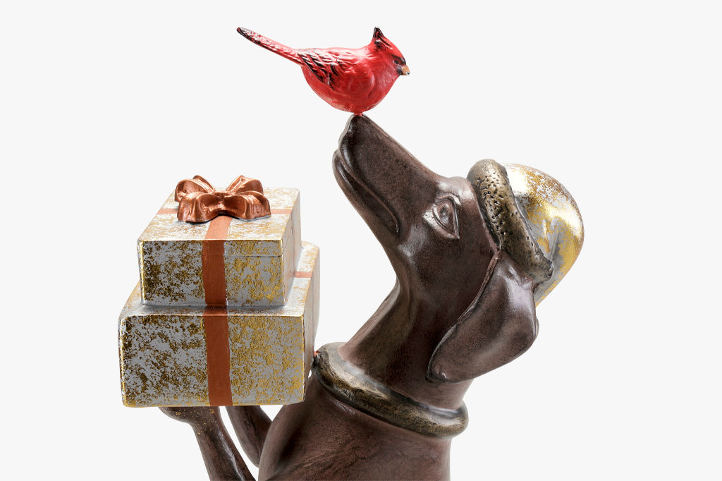Hello, Friend Garden Sculpture features a Dachshund sculpture holding wrapped gifts while a red cardinal perches on its nose. Close up view of gold and silver packages with copper finish bow.