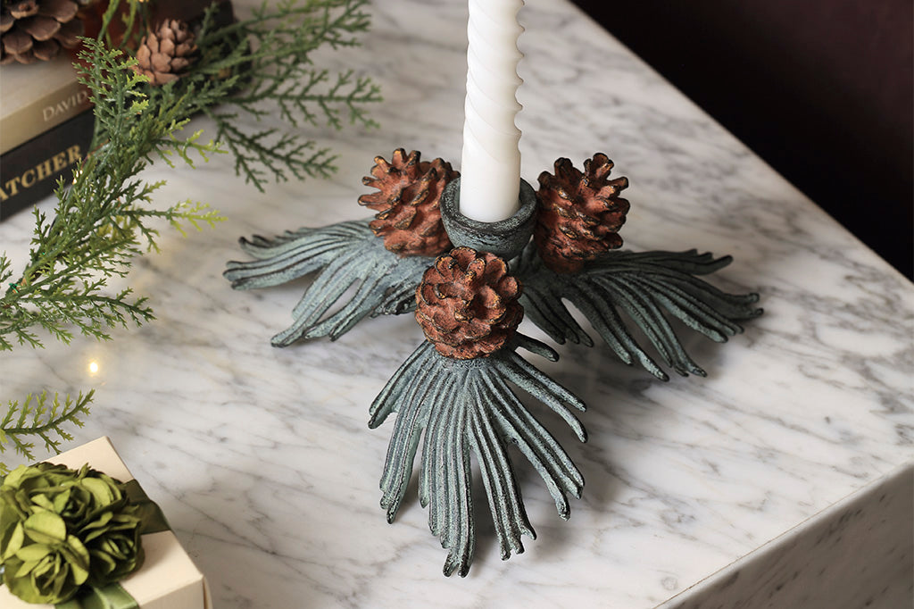 taper candle holder shaped like pineneedle cluster with 3 pinecones shown from top vie on marble counter near faux foliage and wrapped gift