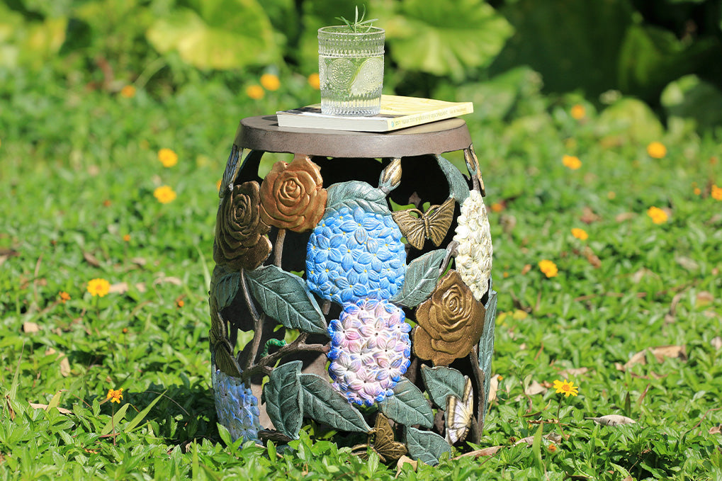 Cast metal garden stool features beautiful roses, butterflies, and the vibrant hues of hydrangeas. Shown on grass outside with book and glass of water on top.