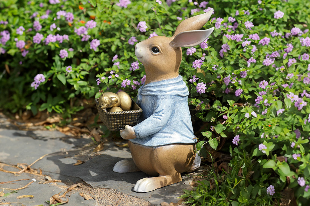 Shown on floral garden path, cast metal sculpture of storybook-style rabbit in a blue shirt, holding a basket of four removable golden eggs. A chick perches on edge of basket to get a closer look.