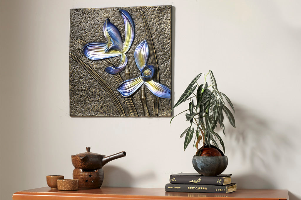 Decorative wall tile with metallic orchid motifs in blue shown hung above a desk potted houseplant and tea set