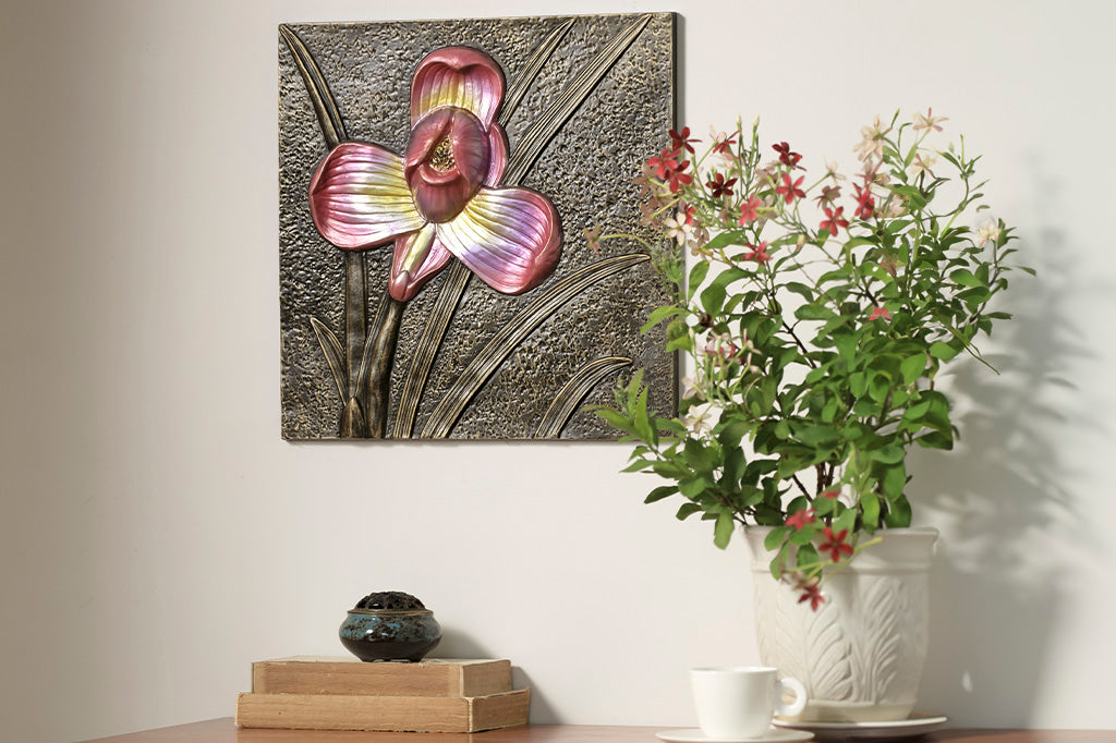 Bronze decorative wall tile with metallic pink/violet orchid motif, shown hung above a desk with books, teacup, and flowerpot