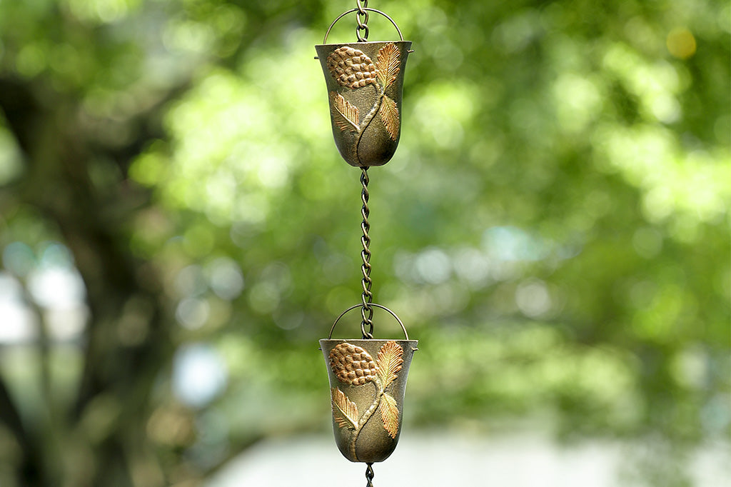 A close up of two cups of a rain chain with 8 cups decorated with pine cone motifs outside on a tree.