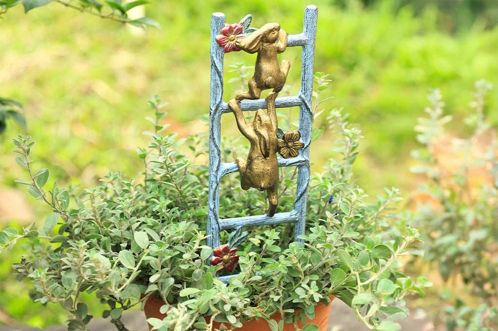 mini planter trellis shown with potted herb in outdoor setting in the sun. Features 2 golden rabbits climbing a ladder with flower vines.