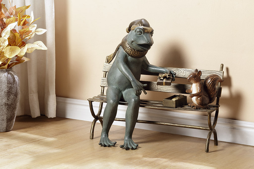 Decorative statue of a frog sharing a box of acorns with a with a squirrel on a park bench, shown on floor in a room setting.