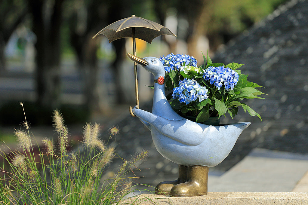 Slate blue goose planter shown holding an umbrella; she wears a flower choker,and rain boots. Planter holds potted hydrangea, placed on concrete stairs by plants.