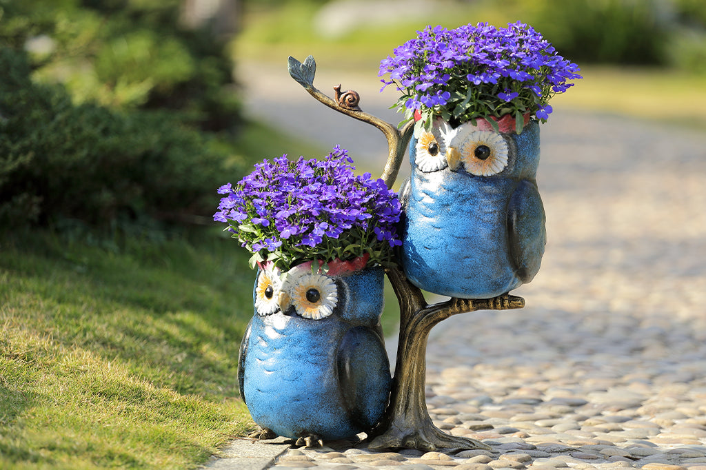 cast metal owl perched on branches planters blue in color holding purple flowering plants set in outdoors