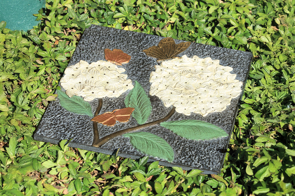 Cast aluminum stepping stone, two white hydrangea with three butterflies fluttering around blooms. Stepping stone set on grass
