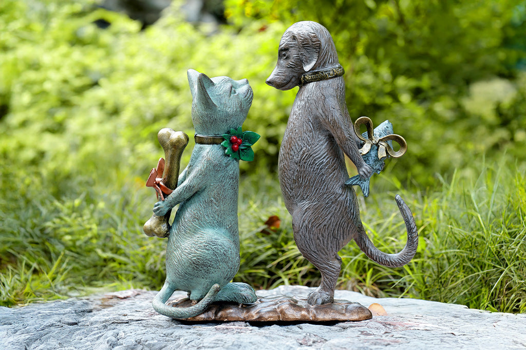 Statue of a cat holding a bone and a dog holding a fish placed in a a garden.