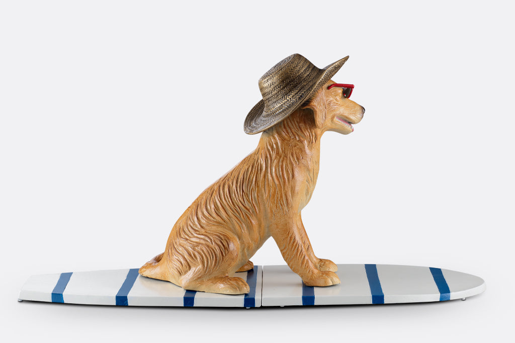 cast metal dog sculpture sat on stripped white and blue surfboard face right. See dogs floppy ears and texture of cast metal straw hat